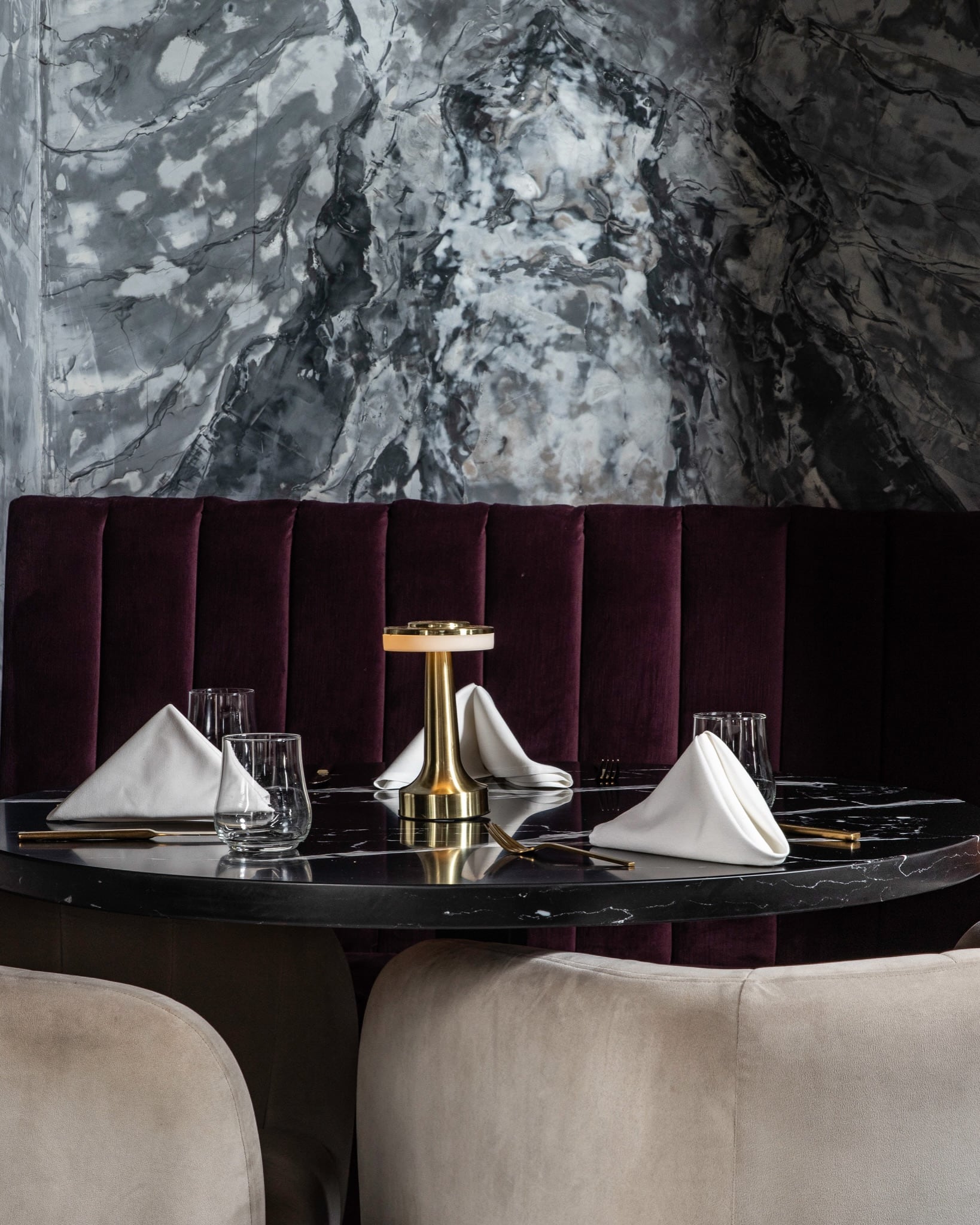 Burgundy channel-tufted banquette with marble wall, black table, cream chairs, and brass table light