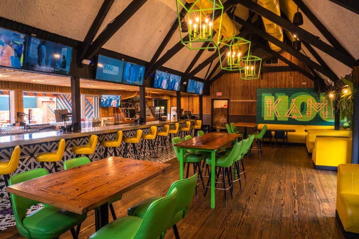 KAMP indoor bar with yellow bar stools, green chairs, wood tables, and yellow booth seating